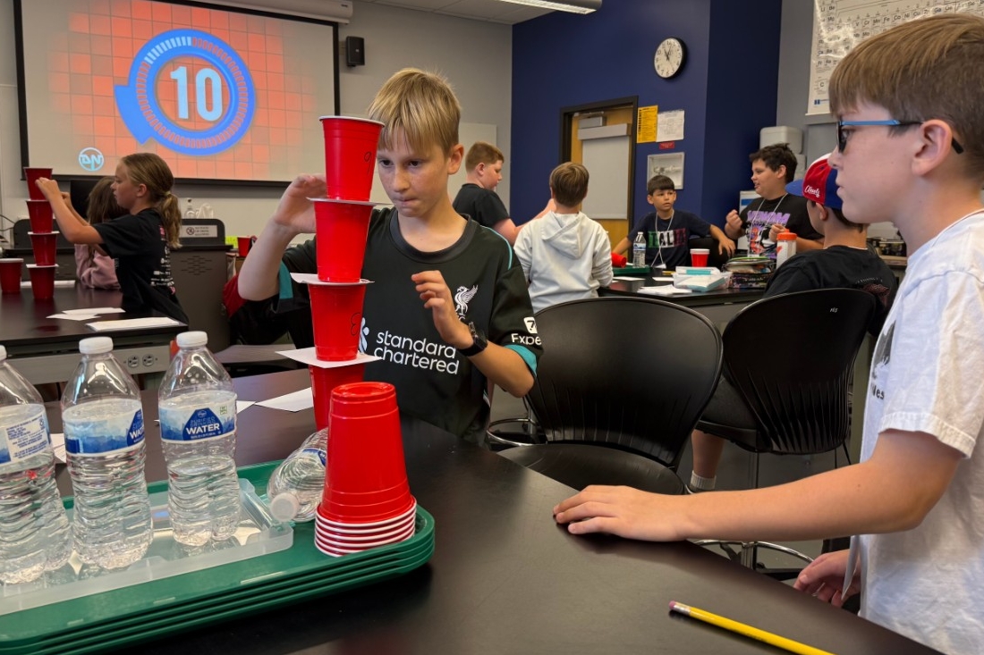 Focused children try to pull paper dividers out from under stacked cups without knocking down the cups.