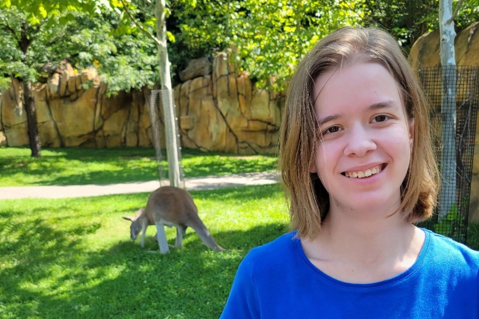 Adrienne Quiroz smiles outside on a sunny day with a kangaroo in the background.