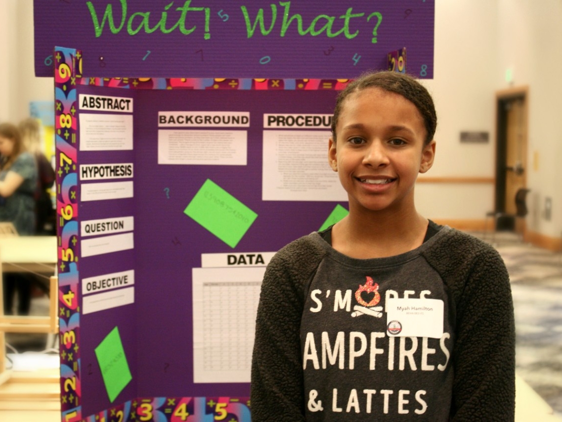 A school-aged student smiling in front of their poster presentation.