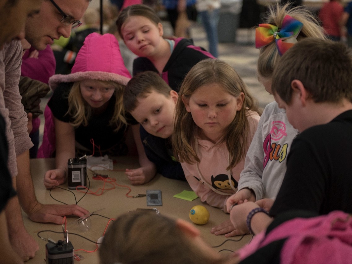 Elementary students gather around and lean in so they can see what will happen in a STEM experiment