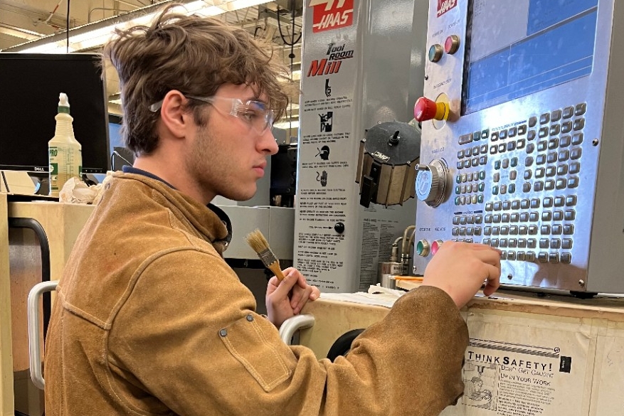 A high school student wearing a protective coat and safety glasses uses a CNC machine to make a custom name tag at Northern Kentucky University.