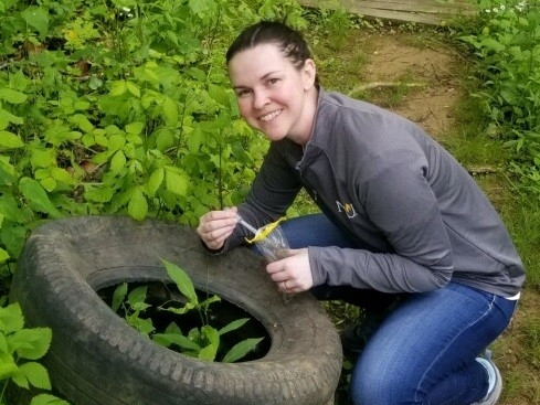 Photo of Dr. Parker, kneeling on a hiking trail, in a grassy area, next to a stack of auto tires.