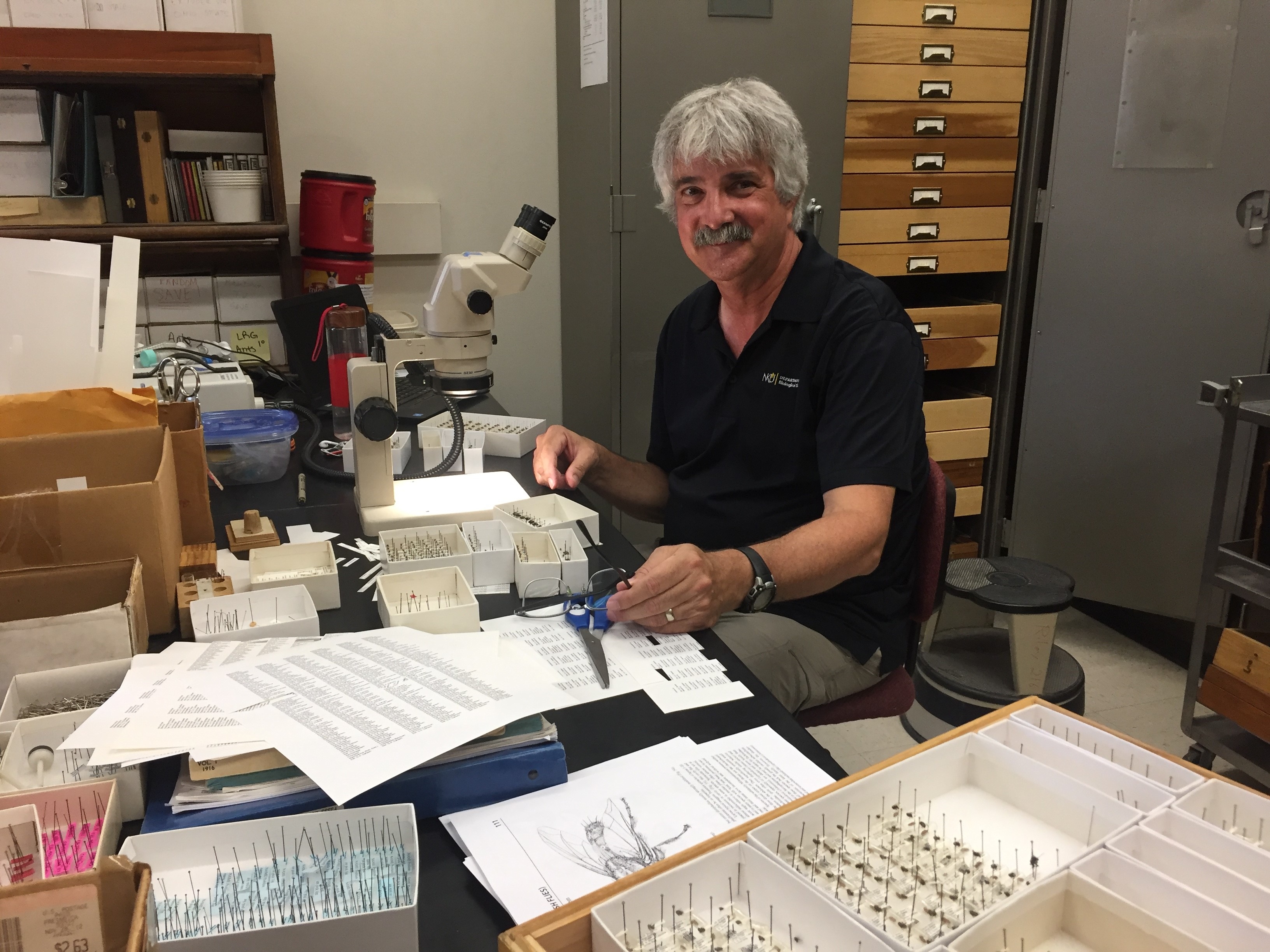 Photo of Dr. Dahlem in a lab. He is sitting in front of a microscrope with boxes of specimen slides next to him on a worktable. 