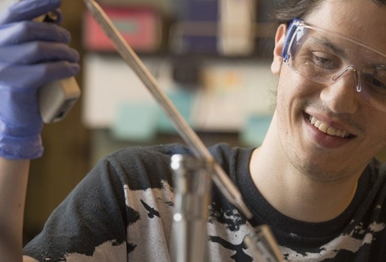 Smiling student wearing protective goggles and gloves, working in lab.