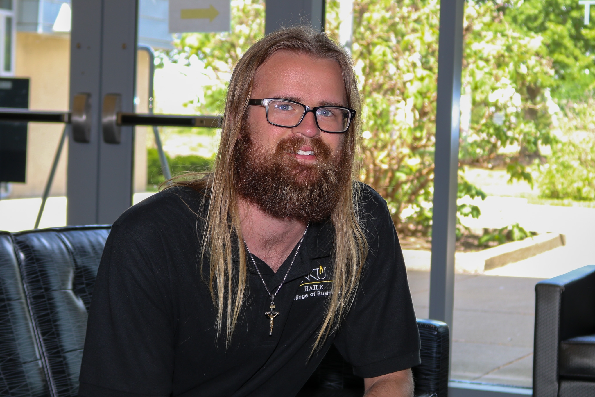 Jacob Albers sits indoors near large windows, smiling at the camera. He has long hair, a full beard, and wears glasses and a black polo with an NKU Haile College of Business logo, with greenery visible outside behind him.
