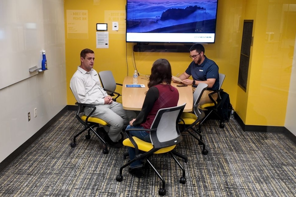 Three individuals sit around a small table in a bright yellow study room, engaged in a focused discussion. One person speaks while the others listen, with a laptop and papers on the table. A wall-mounted screen displays a scenic image, and whiteboards line the walls, creating a collaborative workspace environment.