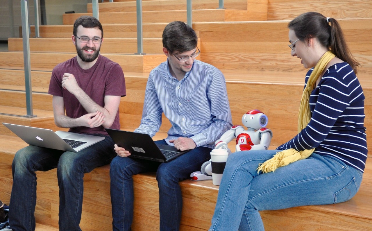 Photo of three students sitting on the steps in Griffin Hall looking at a small bipedal robot