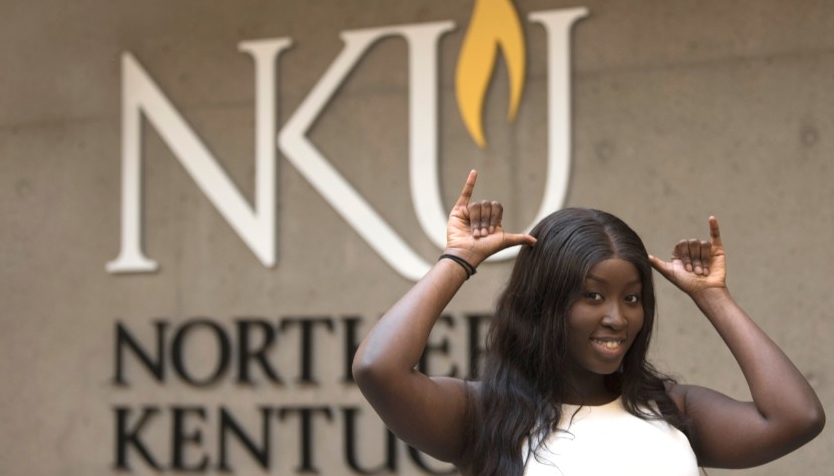 Black female student standing in front of the NKU logo giving a Norse Up