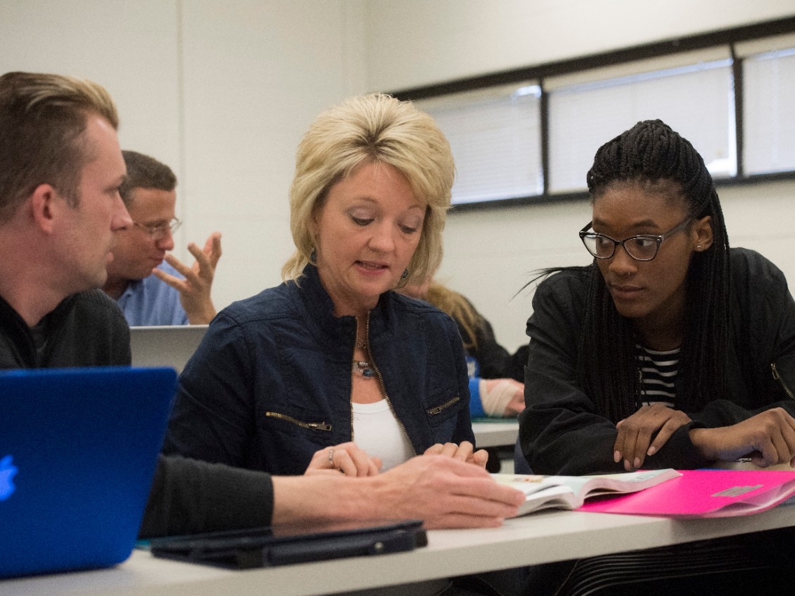 Adult learners sitting in a classroom, looking at a textbook together.